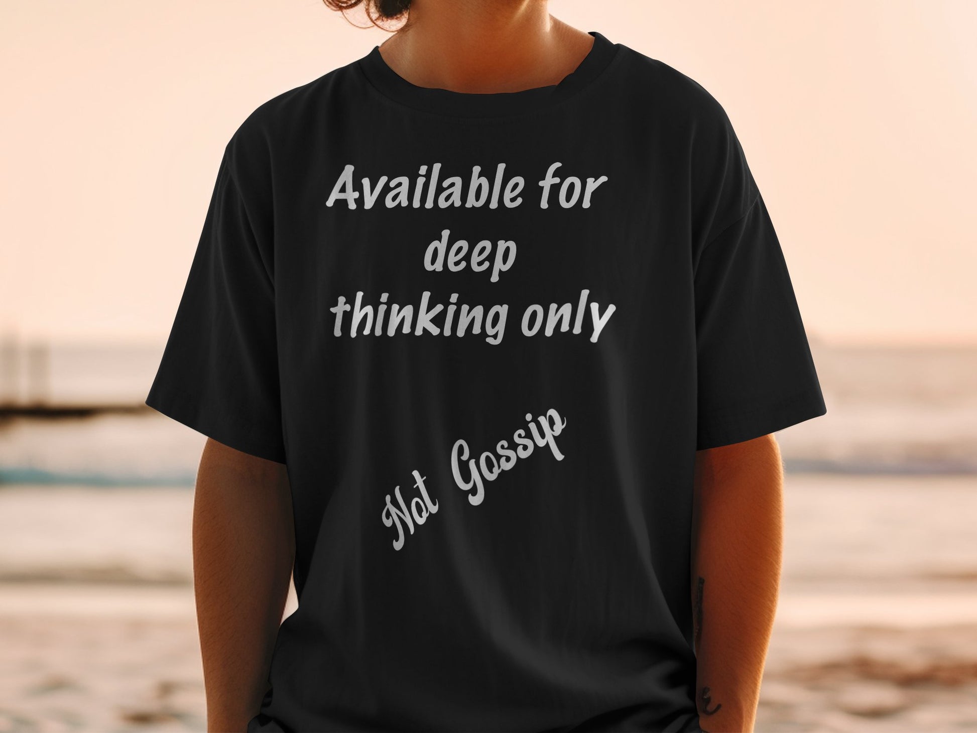 Person wearing a black t-shirt with text 'Available for deep thinking only. Not Gossip' at the beach.